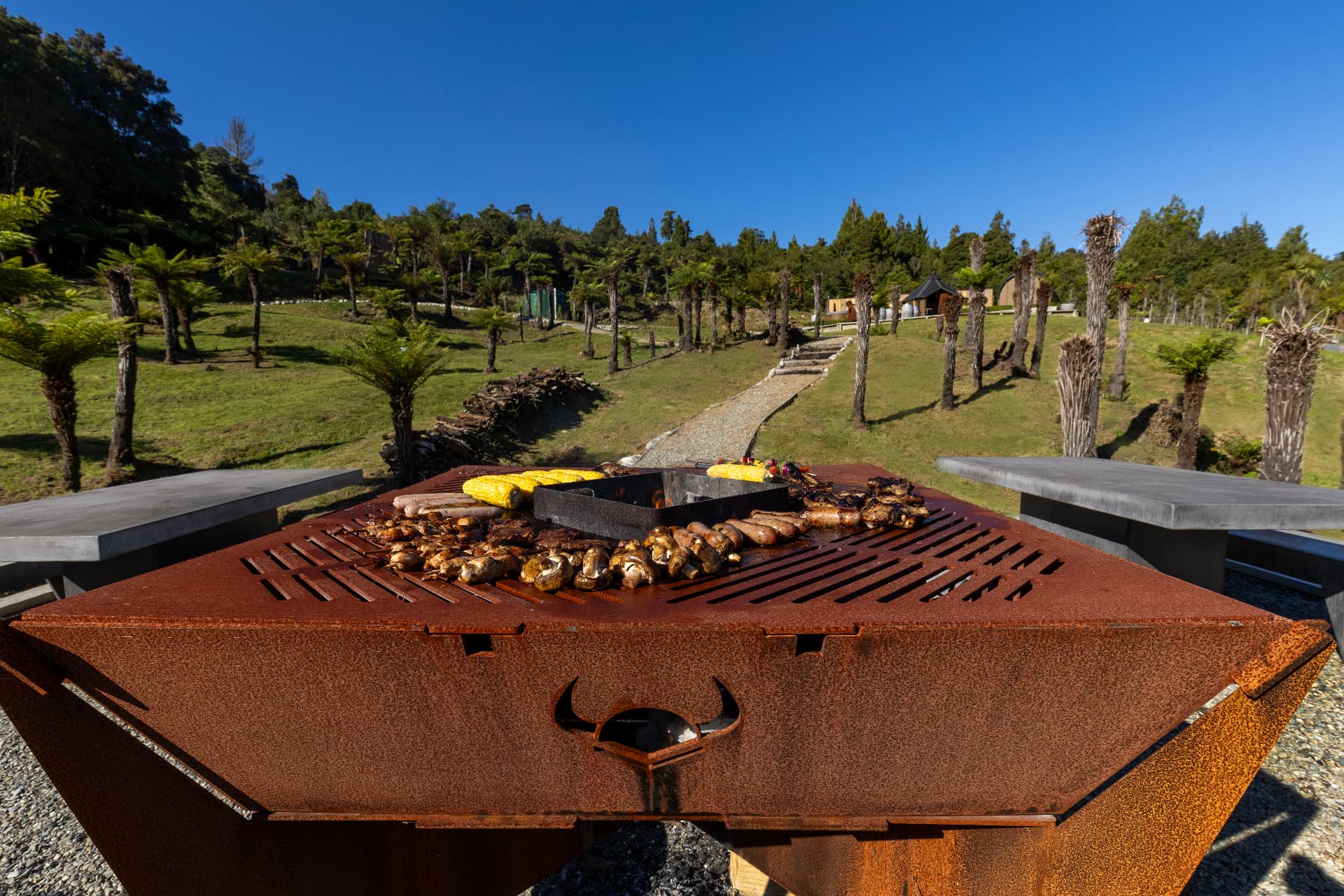 Corten steel barbecue grilling corn, sausages and mushrooms with the barrel cabins and tree ferns beyond